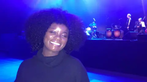 Winnie Ama standing in front of the stage in the Telegraph building in Belfast.  She is smiling at the camera and wearing a black, turtleneck dress.