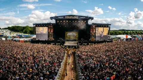 Download Festival A huge aerial shot of the crowd of people at Download Festival. There are thousands packed in front of a large black stage. There are two sides which are barricaded and there is a walkway in the middle.