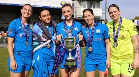 Five Posh Women players standing in a line on the pitch, wearing medals and holding a silver trophy. 