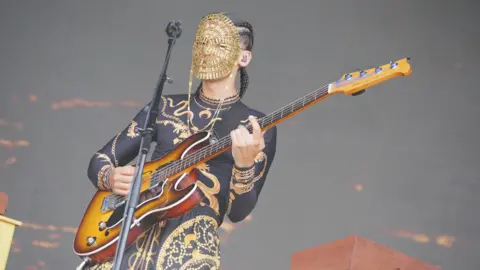 PA Media A performer in a black and golden dress playing the electric guitar in front of a microphone. They are wearing a golden mask, obscuring their face.
