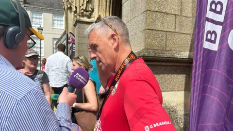 John Askie is standing side-on to the camera as he is interviewed by a man wearing headphones via a microphone that is branded BBC Radio Cornwall.