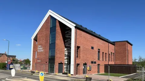 South Warwickshire University NHS Foundation Trust A ground-level view of Ellen Badger Hospital, a modern red brick building with white lettering reading "Ellen Badger Hospital" on the front facade. The building features a tall angular entrance with floor-to-ceiling windows and horizontal louvred panels set into the brickwork. Metal safety barriers are visible in the foreground, with a clear blue sky above.