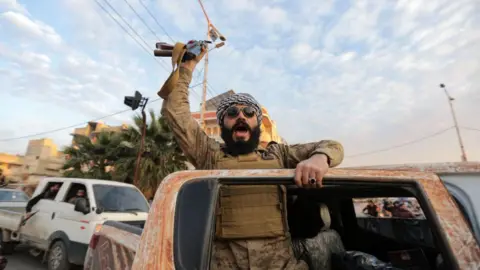 Getty Images A man dressed in all brown military fatigues leans against the door of a rusty red car, weapon raised.