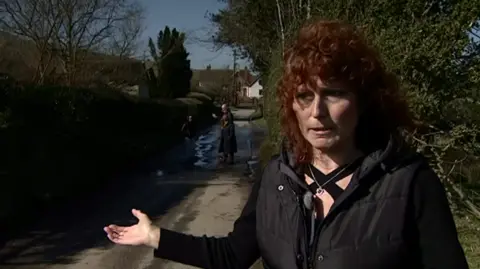 Kim stands on a damaged road with friends in the background. She has her arm out-stretched in anger at the roads. She has curly red hair and is wearing a black top and black jacket.