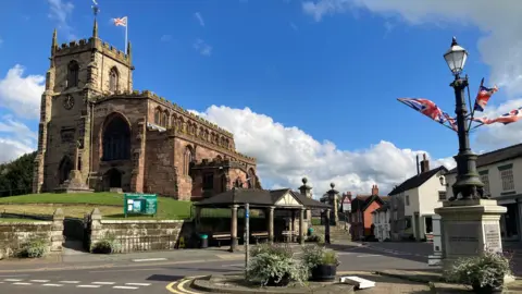 A view of Audlem, which shows the village church on the left, having been taken from across the road opposite a traffic island that has a memorial lamppost with union jacks attached to it.
