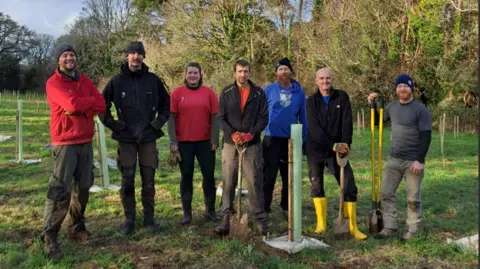 Katie Reynolds A group of people stood on a green field with trees behind them. Three of the people have spades and tools in their hands. All of the people in the photograph are smiling. 