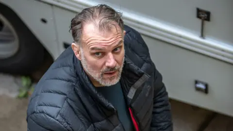 Photo showing Paul Whitehurst looking up at the camera.  He is wearing a navy padded jacket.   He has dark, thinning hair and a grey and white stubbled chin. he wears a puffy blue jacket over a dark green top.