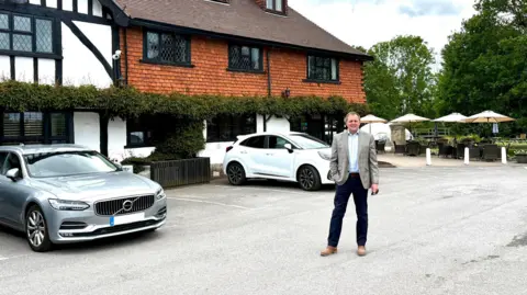 A suited man stands in front of a Tudor-style hotel 