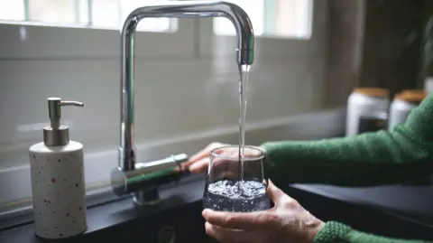 A woman is filling a glass with water from a silver tap. You can only see her hands. She is wearing a green cardigan.