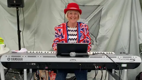 Jean Martyn A woman in a Union Jack blazer and zig zag patterned black and white smiles while sitting at a mini piano. She has short blonde hair in a bog and is wearing a red bowler hat. She is in a tent, with grey material behind her.