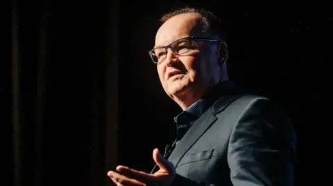 Digital Jersey Tony is speaking on stage, wearing glasses and a dark jacket, gesturing with one hand against a dark background.
