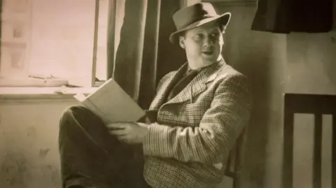 Jeff Towns Dylans Bookstore Dylan Thomas in a black and white photo, sitting on chair beside and open window wearing a Trilby hat and tweed jacket with his legs crossed, a book in his lap and looking up with a slight smile
