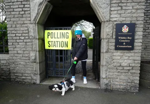 Owen Humphreys / PA Media A black and white dog on a green lead walks alongside a person wearing blue and white bobble hat and black coat as they leave the church polling station.