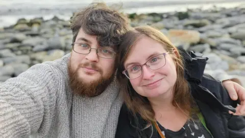 Alyssa Czaja Alyssa and her husband Gareth are sat on a pebble beach, looking at the camera. Gareth is taking the selfie, he is wearing a beige knitted jumper and glasses. Alyssa is wearing a black coat and patterned black and white top, she is also wearing glasses.