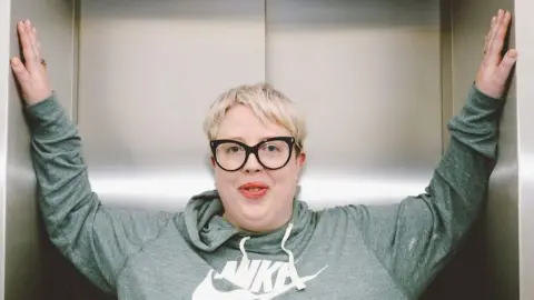 Marea Stamper, alias The Blessed Madonna, pose dans un ascenseur de la BBC New Broadcasting House, 2022. Elle porte un sweat à capuche vert, a des cheveux blonds courts et des lunettes noires.