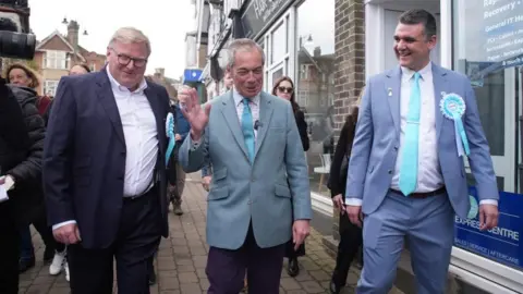 PA Media Nigel Farage wearing a blue-grey blazer and teal tie walks along a street flanked by two men in suits with Reform UK rosettes