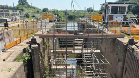 Scaffolding and construction equipment are set up inside a canal lock, with water running through the middle.
