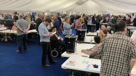 A tent erected inside a sports arena. A blue carpet has been laid down and spotlights illuminate the space. Tables are lined up with people counting votes.