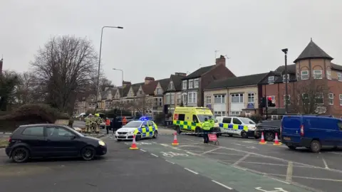 BBC Cones are placed across a road next to traffic lights and a bridge in a city. A sign says 'road closed' and a police car is parked in front of it. A police van can also be seen, along with an ambulance and firefighters on the corner of the street.