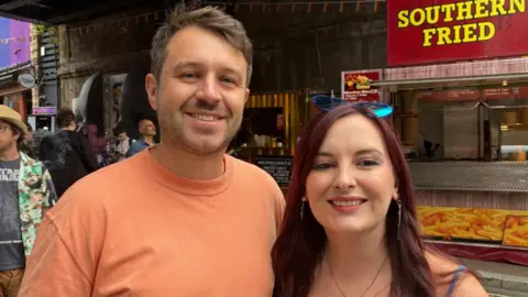 A man and a woman stand next to each other smiling. The man wears an orange T-shirt and has short brown hair and brown stubble. The woman has long brown-red hair and wears sunglasses on top of her head.