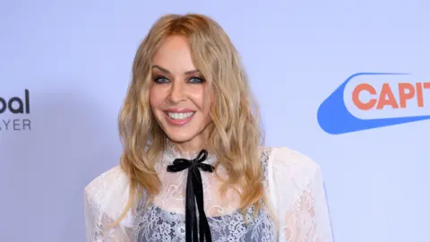 Getty Images Kyle Minogue smiles as she stands in front of a Capital Radio awards background. She has long curly blonde hair coming down below her shoulders, a white lacy top with a black neck tie