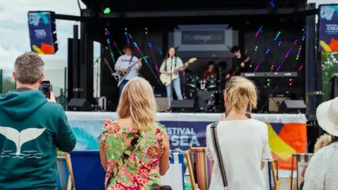 North East Lincolnshire Council A band, which includes three guitarists and a drummer, perform on a stage as people, with their backs turned to the camera, watch the concert.
