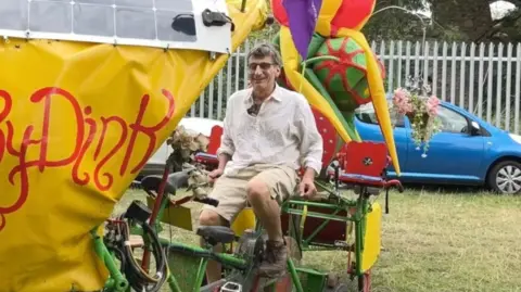 A man sat on the rinky dink - it is a pedal powered machine. He is looking at the camera and smiling.