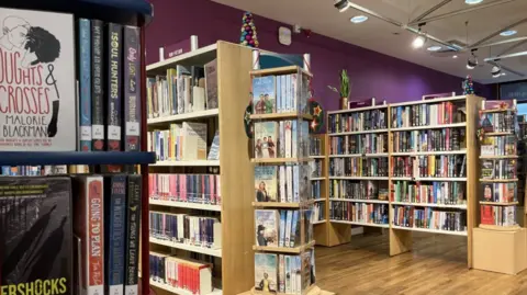 Several wooden shelving units are filled with books. The walls are painted purple and the flooring is wood.