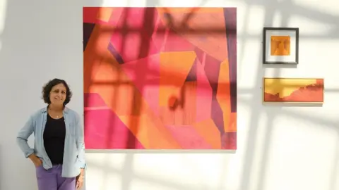 Irina Mackie Suminder Virk stands next to paintings displayed on a gallery wall.