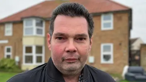 Alex Dunlop/BBC Phillip Shaw smiles at the camera. He has dark, short hair. He is wearing a black coat with a checked shirt underneath. He is standing in front of a house, which is out of focus.