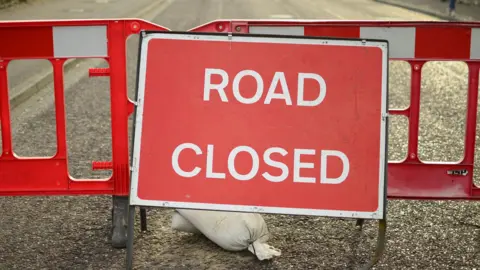 A stock image of a red plastic barrier with a road closed sign in front of it which is on a section of road where a pavement is just visible at the side.