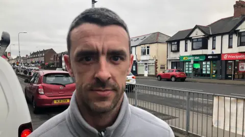 Joshua Riley looking at the camera. He is stood on Newport Road with cars behind him. He is wearing a grey fleece. 