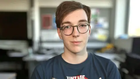 Martin Giles/BBC Mac also pictured in a classroom. The background is out of focus. He has brown hair brushed to one side. He is wearing a blue top and brown glasses with round frames.