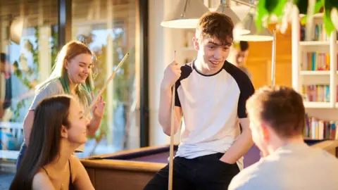 sturti / Getty Images A teenage boy with a pool cue sits in a brightly lit room talking to two friends