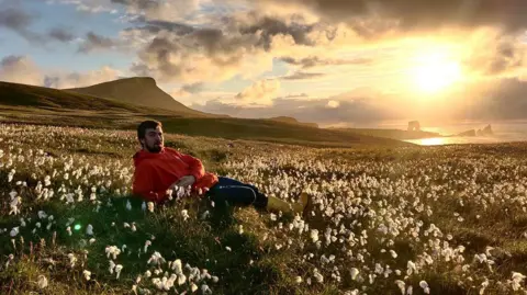 Robert Smith Man with beard and wearing red hoodie, dark trousers and yellow wellies, lying amid flowers in a field with hands clasped, and island coastline in background and sea, with sun glowing brightly in the sky.