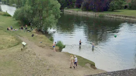 People sitting on the grassy bank of River Thames at Wallingford. A couple of people are in the water, which is up to their knees.