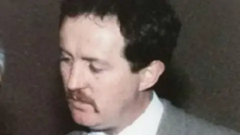 A PSNI handout image of John Devine. He has dark hair and a moustache. He is wearing a grey suit and white shire. 