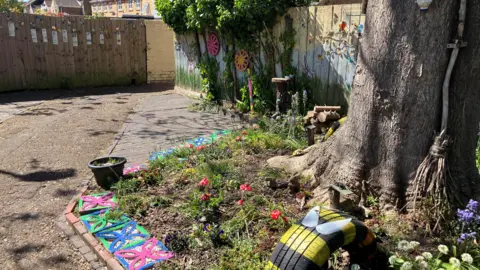 Shariqua Ahmed/BBC A big tree in the street with decorations around it and a fence panel behind it with colourful murals.