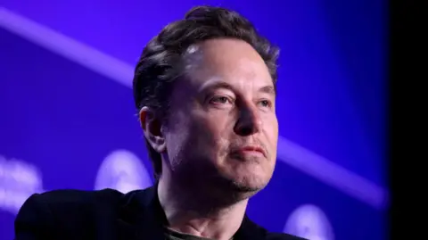 Reuters Elon Musk stares into the distance as his face is lit on stage, with a screen behind him, at a conference