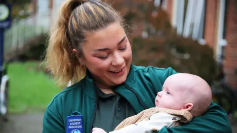 University of Worcester A student paramedic wit light brown hair tied in a ponytail. She is smiling down at a baby in her arms. She is wearing a teal t-shirt and jacket. On the jacket it reads "university student paramedic".
