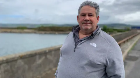A man with grey hair in a grey fleecy top stands in front of the harbour wall in Stranraer