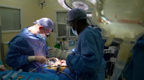 Two surgeons, wearing blue overalls and face masks, are operating on a person in a hospital. They are surrounded by medical equipment. 