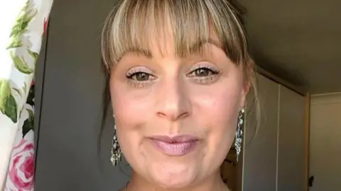 Sarah Albone: a woman with short light brown hair wearing long silver earrings.
