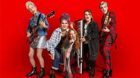 BBC/Drama Republic/Julia Kennedy Five middle aged women wearing punk rock outfits, eahc holding a different musical instruments and standing against a red background.