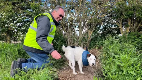 Amanda White/BBC Steve Priestman of Drone SAR for lost dogs UK pictured with Krakka. He is wearing a yellow hi-vis jacket and is smiling. Krakka is white with brown and black markings. 