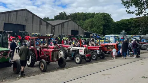 Rows of vintage tractors on display in a farmyard. There are two large warehouse style buildings behind and trees. The floor is concrete and tarmac.