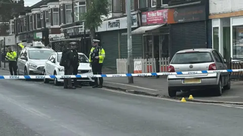 BBC A silver VW Polo on the pavement in front of a row of shops. There is a police cordon in place, with tape across the road.