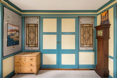 Historic England Archive One of the rooms inside Sharland House. The walls and double doors are painted turquoise and a pale yellow. There is a seascape hanging on a wall and underneath it is a low pine chest of drawers and opposite is a grandfather clock. There are two leaded panels either side of the doors which are ornate, featuring a diamond shape in a frame. 