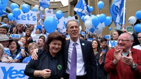 Getty Images A smiling man in a suit, shirt and tie has his arm round the should over a smiling woman as they stand in front of cheering crowds