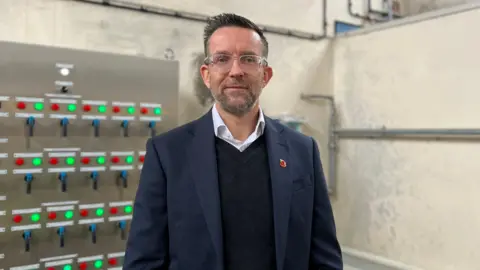 Steve is wearing a white shirt underneath a black v neck jumper. He is wearing a navy blue blazer on top. He has short dark brown hair on his head and has dark brown facial stubble and is wearing frameless glasses.